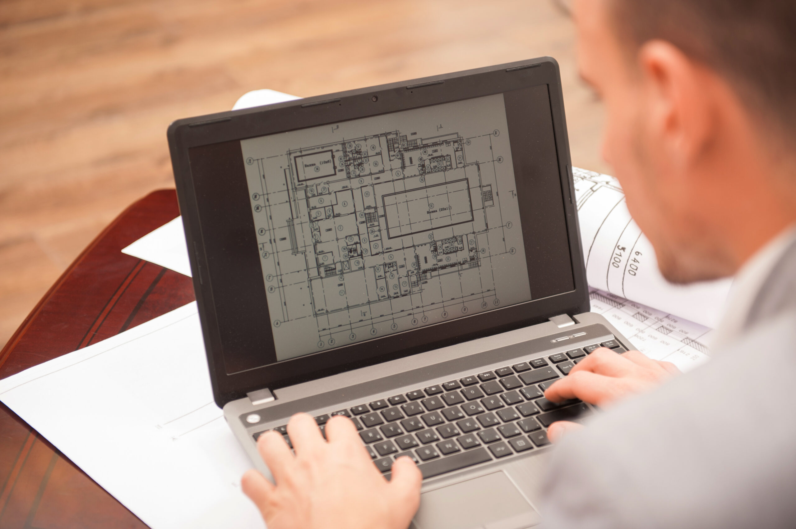 Close-up portrait of laptop with blueprints, architect sitting from behind working on architectural plan, interior shot